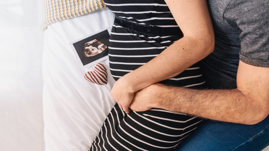 Mujer embarazada es sostenida por su pareja. Ambos están sentados en la cama, a un lado de ellos hay una fotografía de ultrasonido de su hijo.